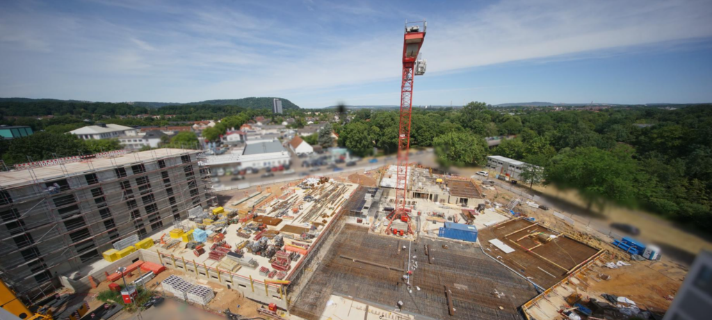 Blick von oben auf die Baustelle am Husarenweg mit Kran, Rohbauflächen und umliegender Stadtlandschaft.